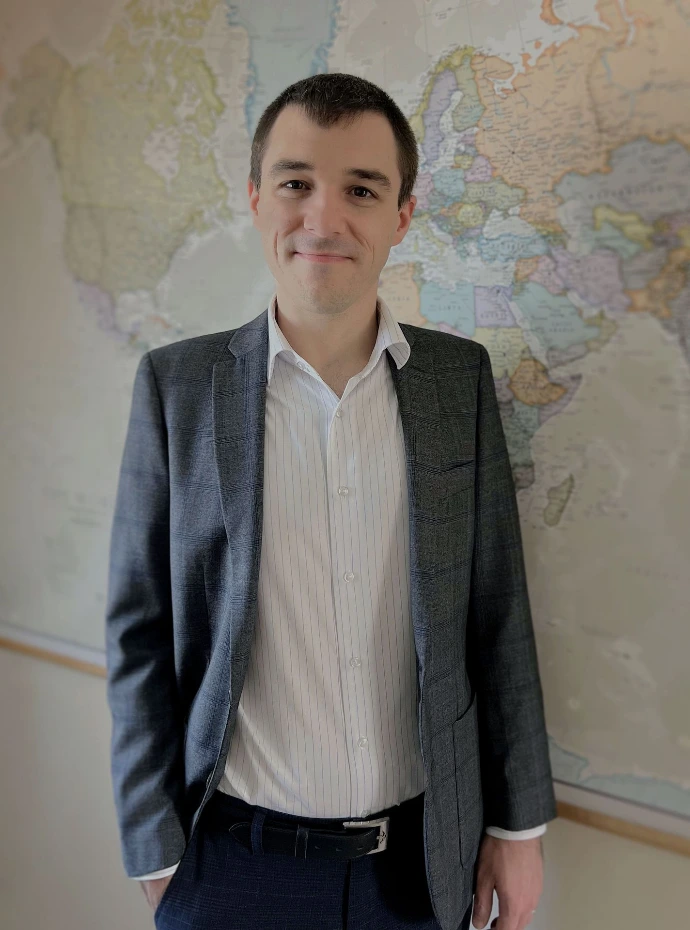 Image of Tone Veca, smiling, standing in front of a world map hung on a wall. Tone is wearing a white business shirt with blue suit jacket.