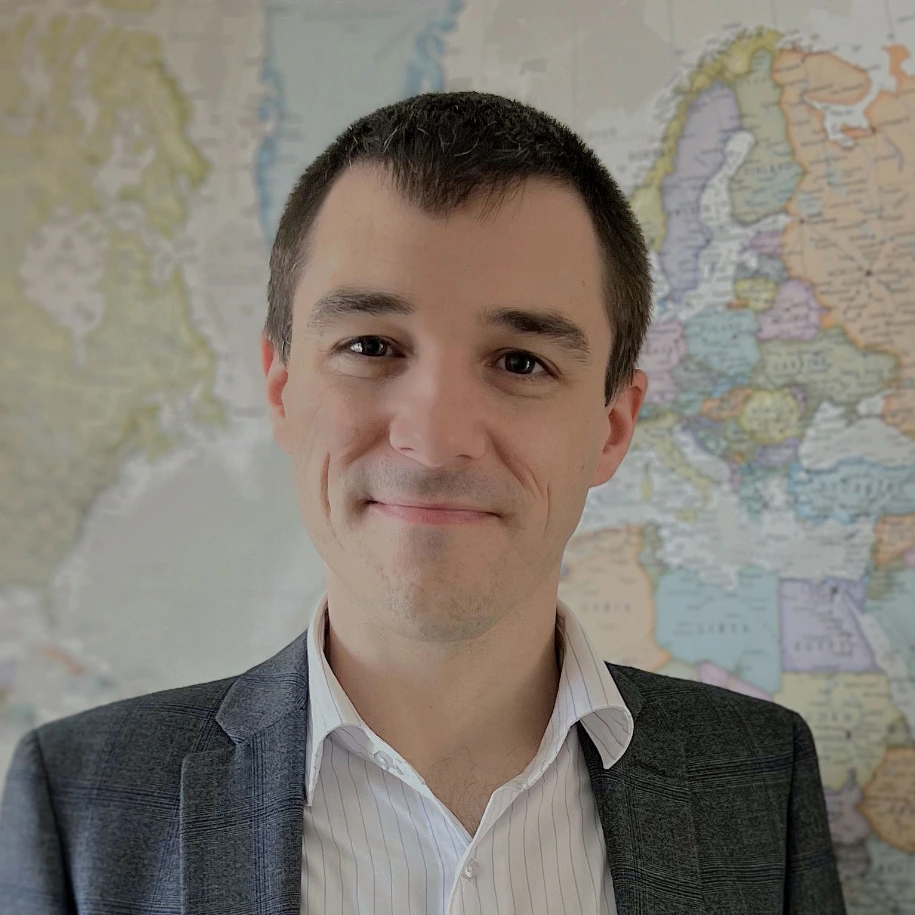Image of Tone Veca, smiling, standing in front of a world map hung on a wall. Tone is wearing a white business shirt with blue suit jacket.
